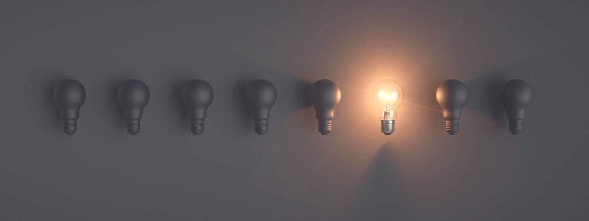 Einzelne leuchtende Glühbirne zwischen sieben nicht leuchtenden Lampen an einer grauen Wand.
