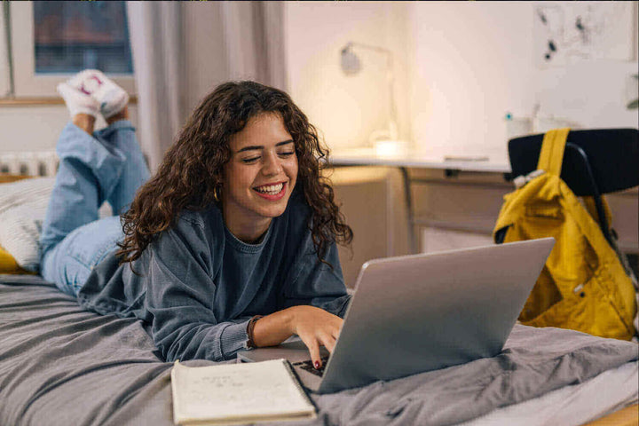 Junge Frau liegt auf dem Bett und nutzt einen Laptop, ideal für Tipps zum Thema: Wie beleuchtet man ein Jugendzimmer richtig?