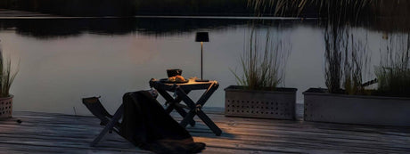 Akku Tischleuchte auf einem Tisch am Ufer mit reflektierendem Wasser, schafft eine gemütliche Atmosphäre im Freien.