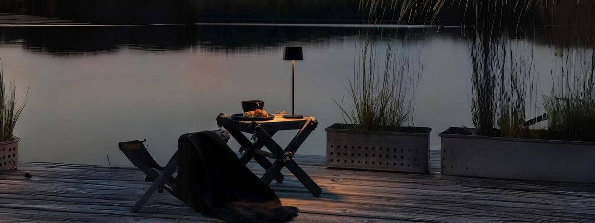 Akku Tischleuchte auf einem Tisch am Ufer mit reflektierendem Wasser, schafft eine gemütliche Atmosphäre im Freien.