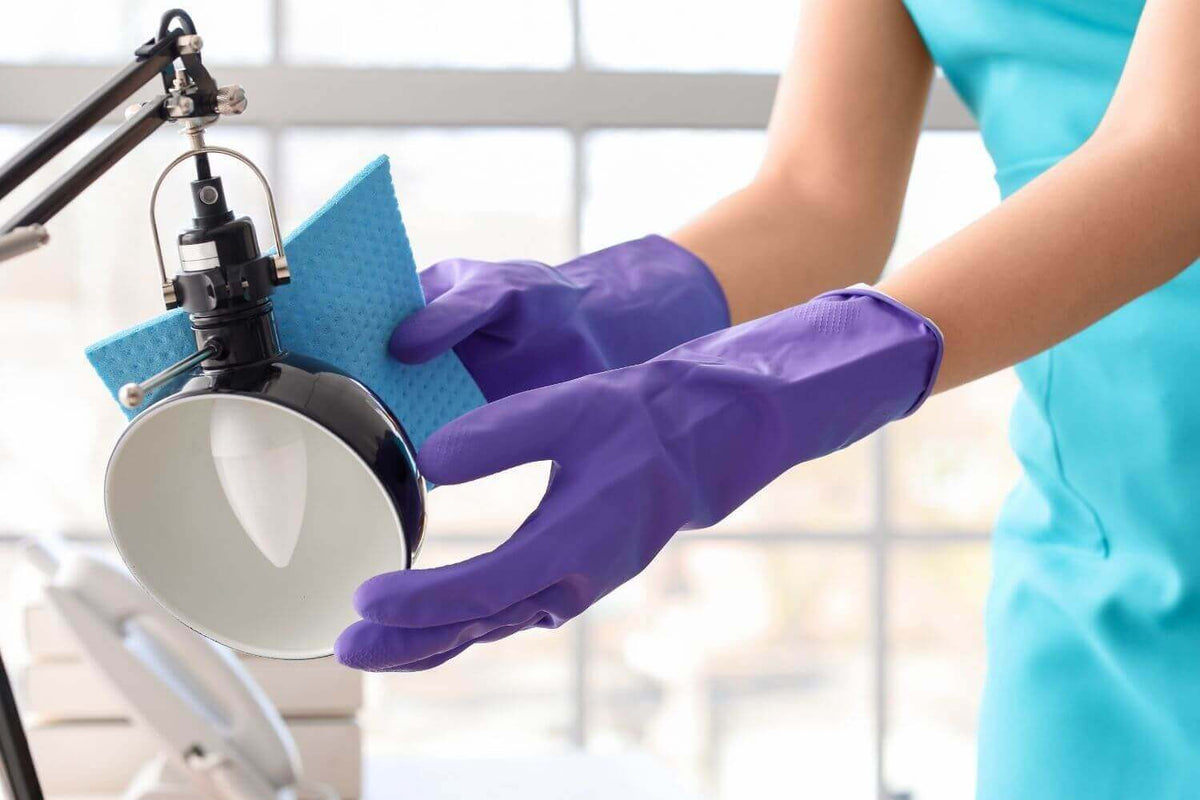 Person mit lila Handschuhen reinigt einen Lampenschirm mit einem blauen Tuch.