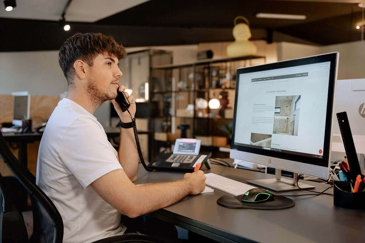 Mann im Büro am Telefon, arbeitet an Computer mit Monitor und Bürobedarf.