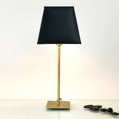 Lampada da tavolo Mattia quadrata in ferro oro-nero con paralume nero e struttura dorata su tavolo liscio.