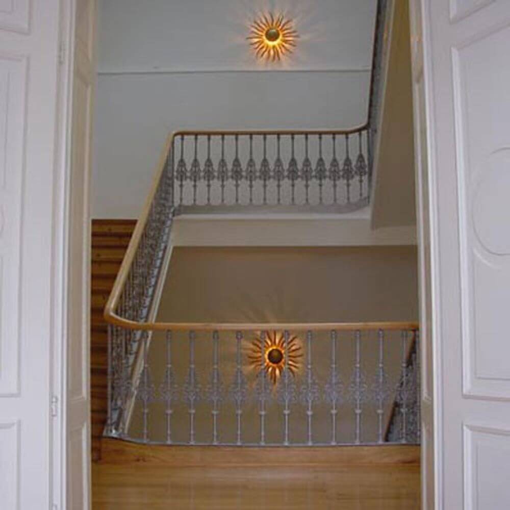 s.luce Diator XL handgeschmiedete Sonne Rost- & Goldfarben an einer Treppe mit elegantem Geländer.