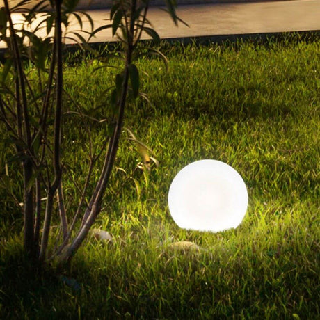 Bolla Garten Außenkugel Ø 30cm Weiß leuchtet sanft auf einer grünen Wiese, umgeben von Pflanzen.