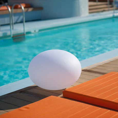 La lampe design à LED à batterie Flatball XXS avec contrôle par application à côté d'une piscine sur une terrasse en bois.
