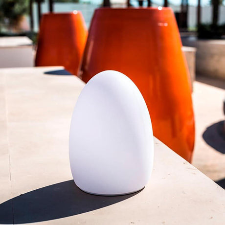 Accu LED tafellamp Egg met app-bediening op een tafel buiten, omringd door kleurrijke meubels.