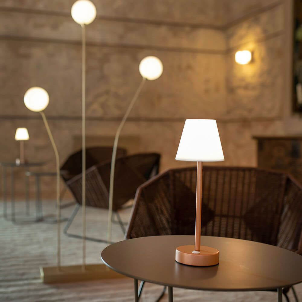 Akku LED-Tischleuchte Lola auf einem Tisch, umgeben von stilvollen Sitzgelegenheiten in elegantem Ambiente.