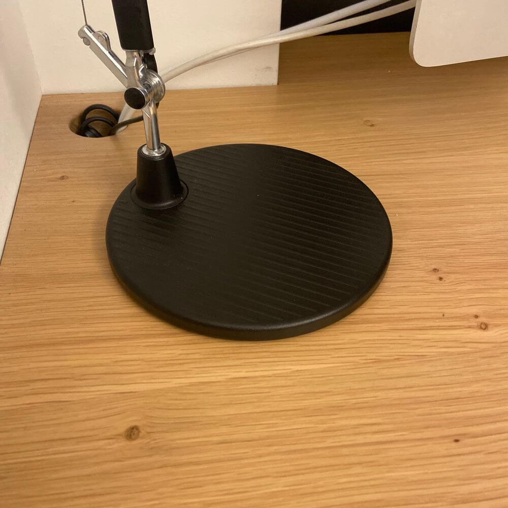 Schwarzer, runder Lampenfuß der Artemide Tolomeo Micro Tischlampe auf einem Holzschreibtisch.