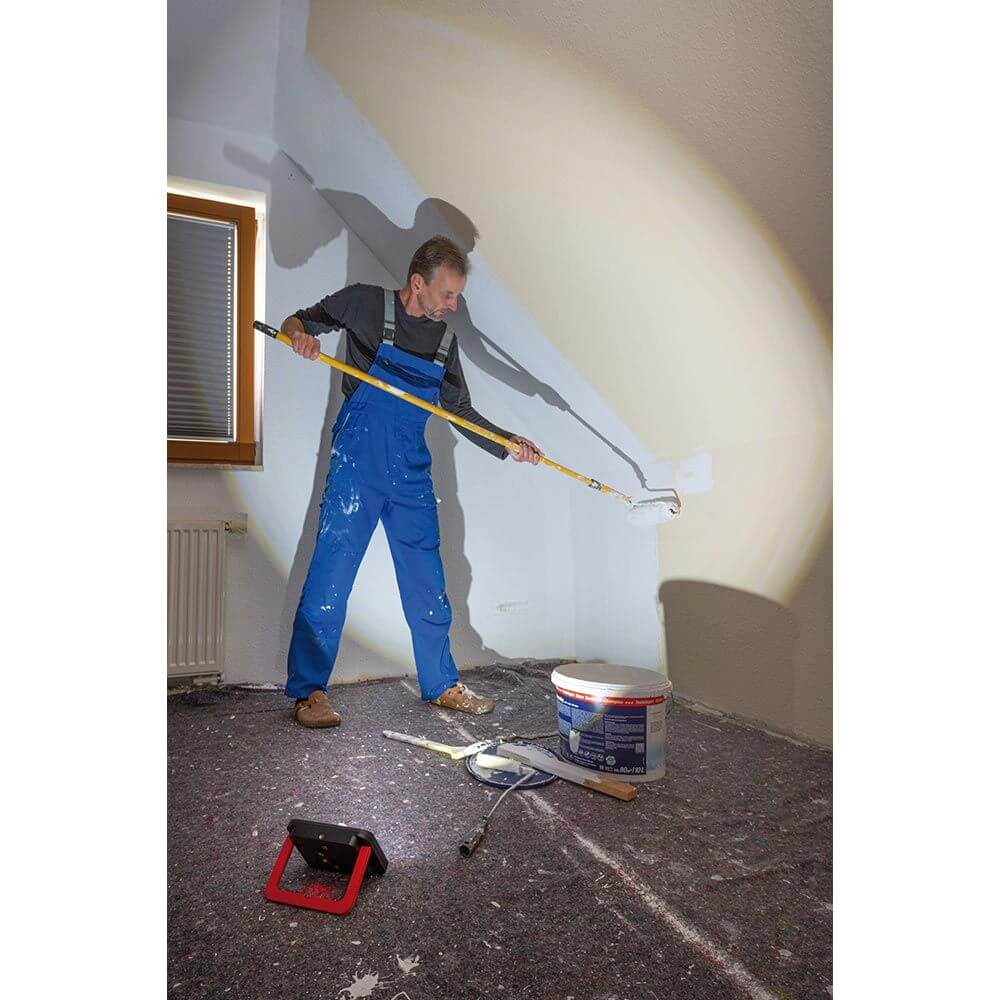 Mann streicht eine Wand mit einer Farbrolle und einer LED Arbeitslampe im Hintergrund.