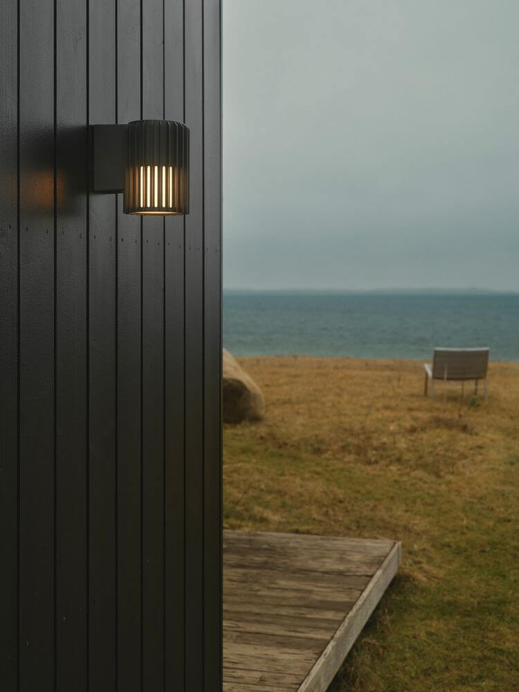 Außenwandlampe Aludra Seaside IP54 an einer schwarzen Wand im Außenbereich mit Blick auf das Wasser.