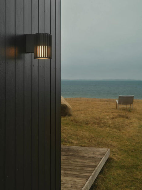 Utendørs vegglampe Aludra Seaside IP54 på en svart vegg utendørs med utsikt over vannet.