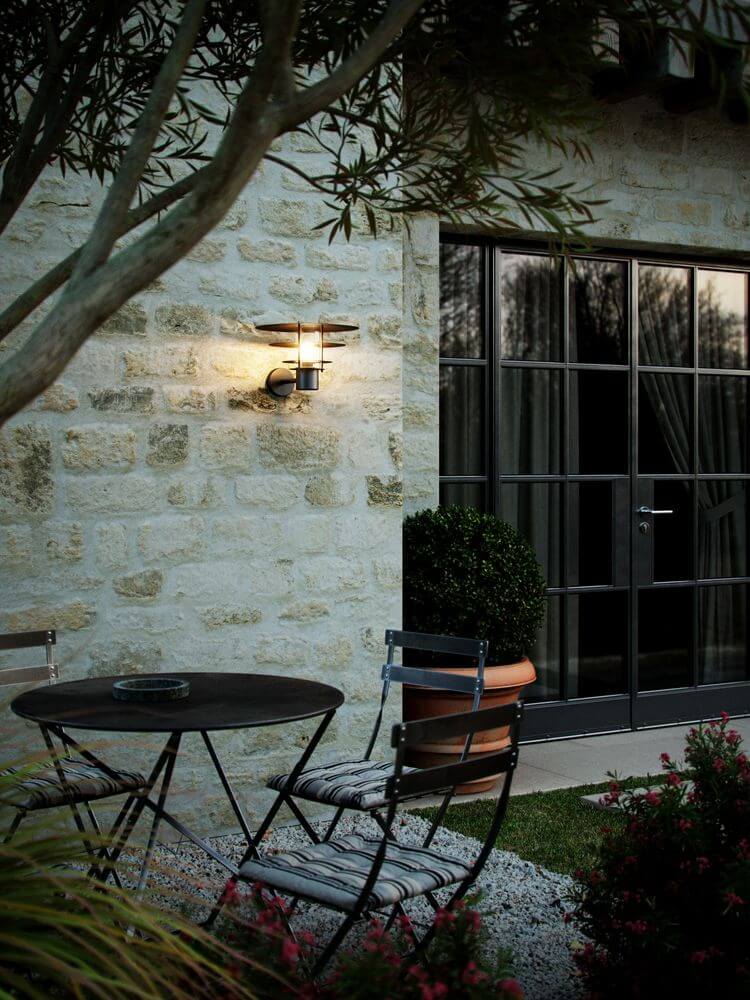 Außenwandlampe Bastia 35 IP54 an einer Steinwand neben einem Tisch und Stühlen im Garten.