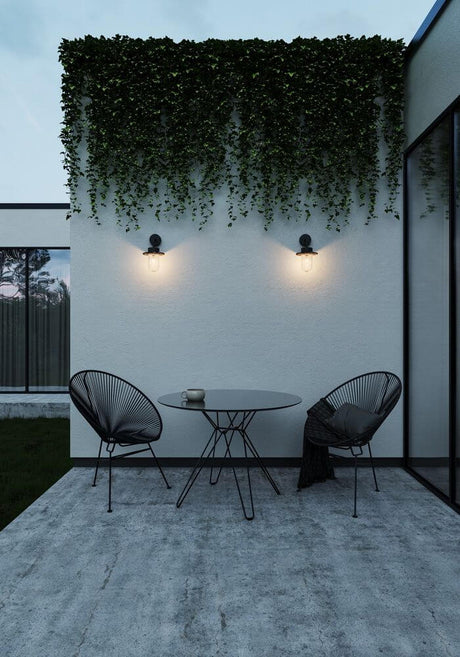 Außenwandlampe Januka IP54 montiert an einer Wand mit Pflanzen, stilvoller Außenbereich mit Tisch und Stühlen.