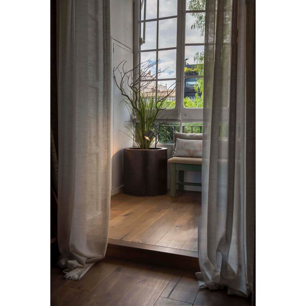 Innenansicht mit Pflanzen und Sitzbank vor einem Fenster, umgeben von Vorhängen.