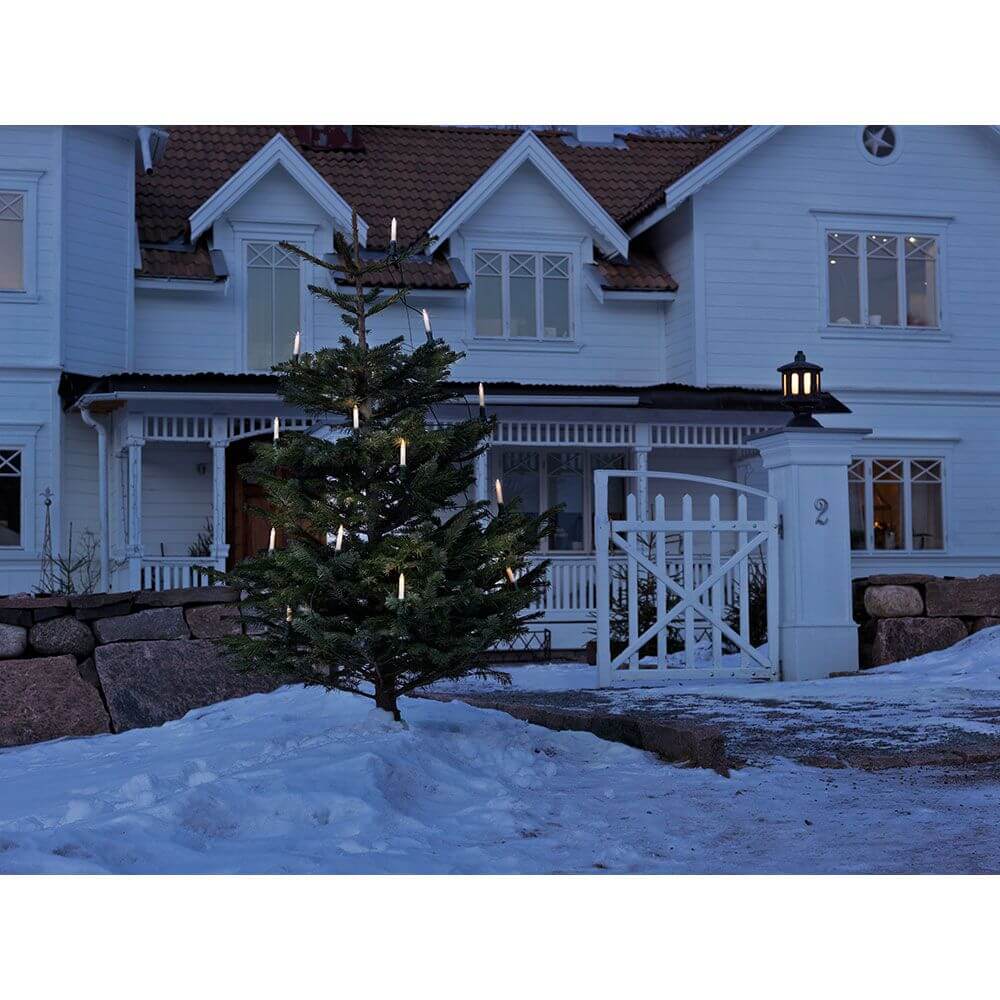 Baumkette Schaftkerzen teilbarer Stecker 16 klare Birnen IP44 beleuchtet einen Weihnachtsbaum im Schnee vor einem weißen Haus.