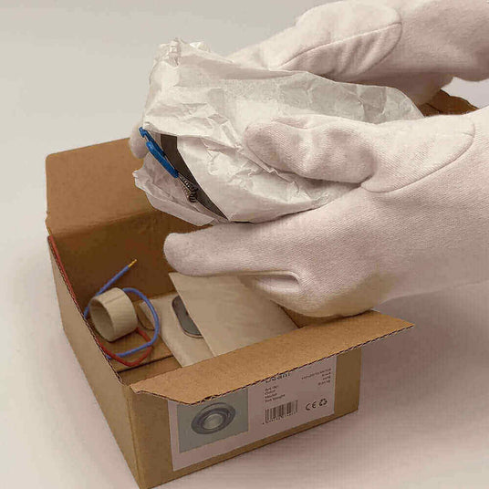 Hand mit weißen Handschuhen hält ein eingewickeltes Paket aus einer Box, die Elektroteile enthält.