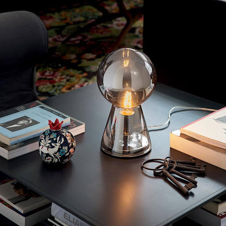 Lampa stołowa Birillo z ręcznie dmuchanego szkła 30 cm dymiona na stole z różnymi książkami i dekoracyjną misą.