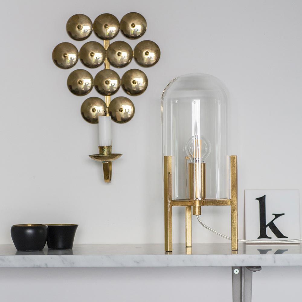 Smokey bordlampe med glasskuppel ved siden av gullfarget vegglampe på en lys hylle, stilfullt oppholdsrom.