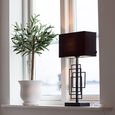 Lampada da tavolo Paragon M con paralume in tessuto nero accanto a una pianta nella finestra, atmosfera moderna e artistica.