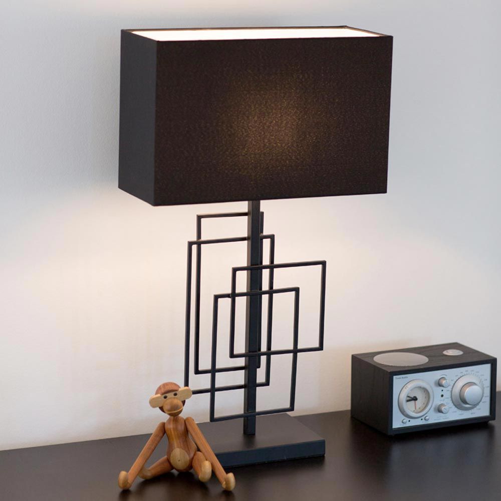 Moderne Tischleuchte Paragon L mit schwarzem Stoffschirm und grafischem Metallrahmen auf einem Tisch.