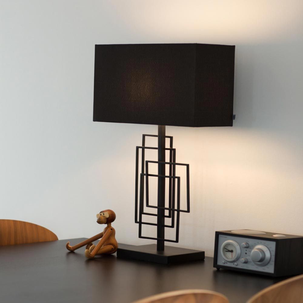 Tischleuchte Paragon L mit schwarzem Stoffschirm und modernem Metallrahmen auf einem Tisch, daneben eine Holzfigur.