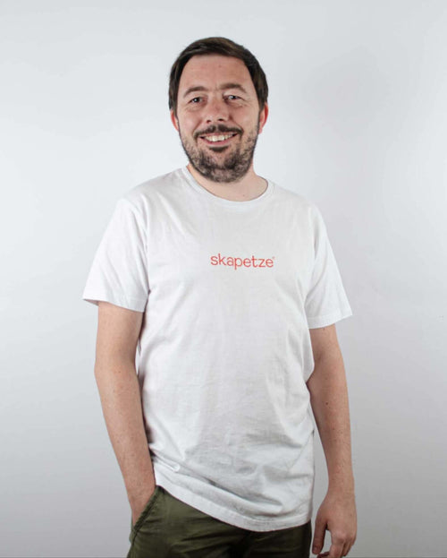 Lächelnder Mann in weißem T-Shirt mit rotem Aufdruck "skapetze", vor neutralem Hintergrund.