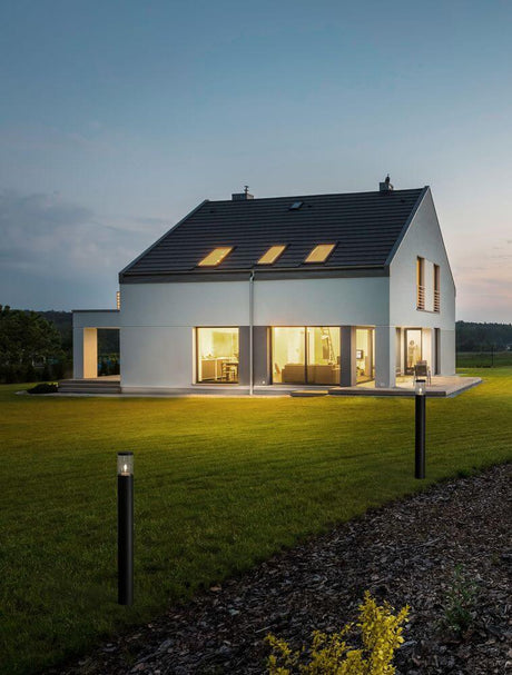 Applique d’extérieur de chemin Cetera noire IP54 à côté d’une maison moderne dans la lumière du soir sur une prairie verdoyante.