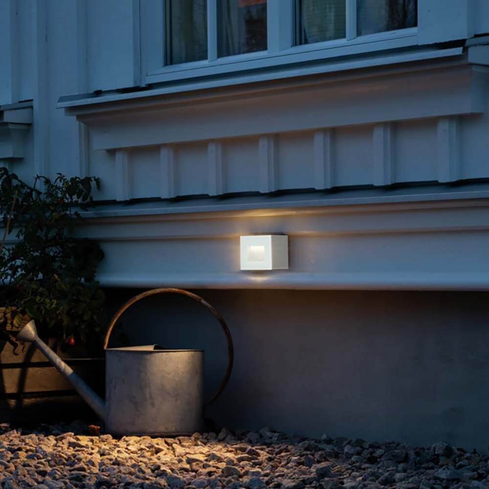 Chieri LED Außen Wandleuchte an einer Hauswand im Freien, in warmem Licht, umgeben von Gartenmaterialien.