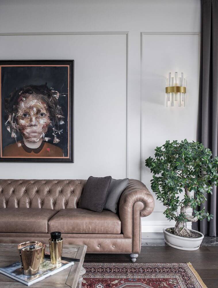 Crown Wandleuchte Gold-Satiniert neben einem braunen Chesterfield-Sofa in einem stilvollen Wohnraum.