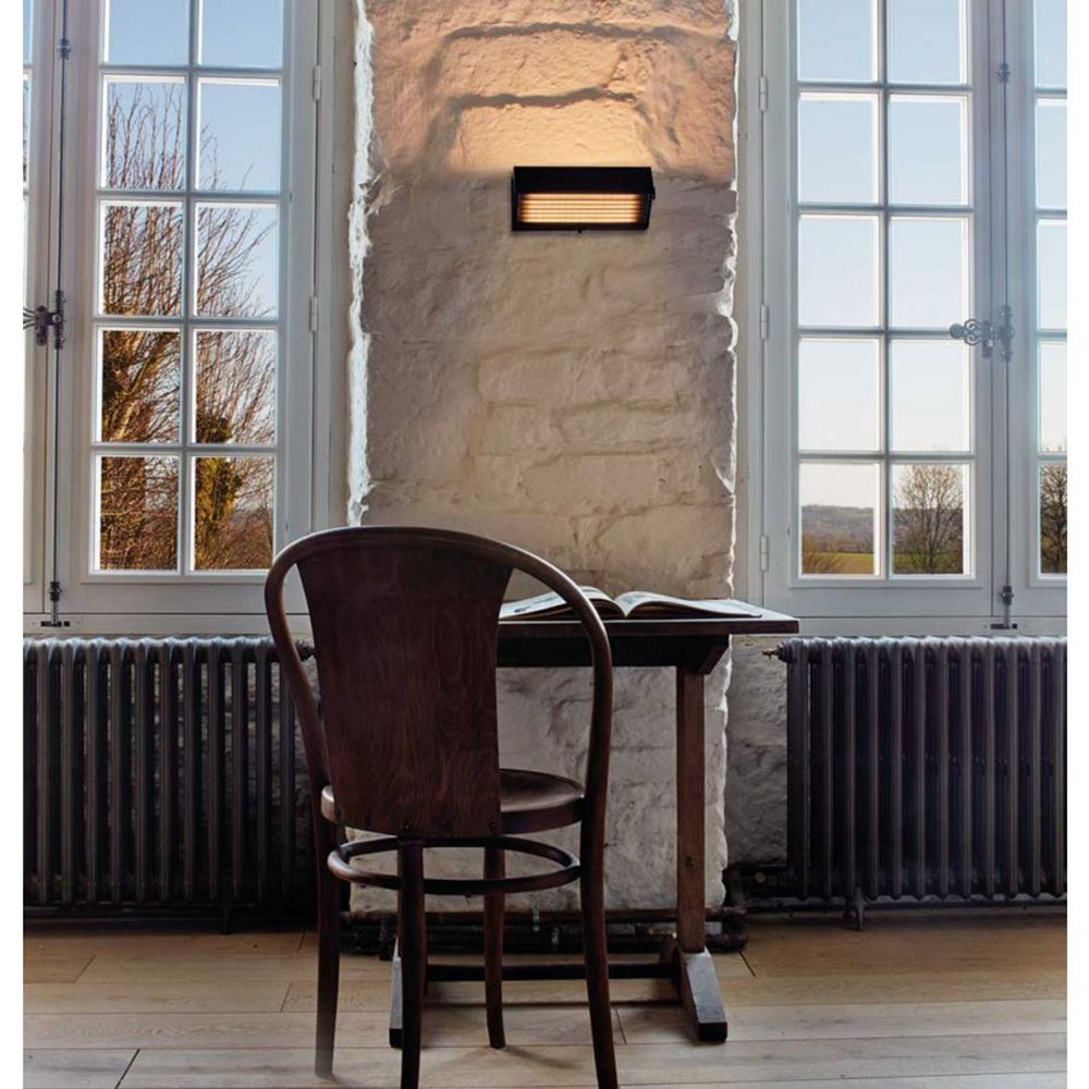 DCWéditions Biny Box 3 Wandleuchte an einer Steinwand mit Fenster und Holzstuhl, schafft eine gemütliche Beleuchtung.