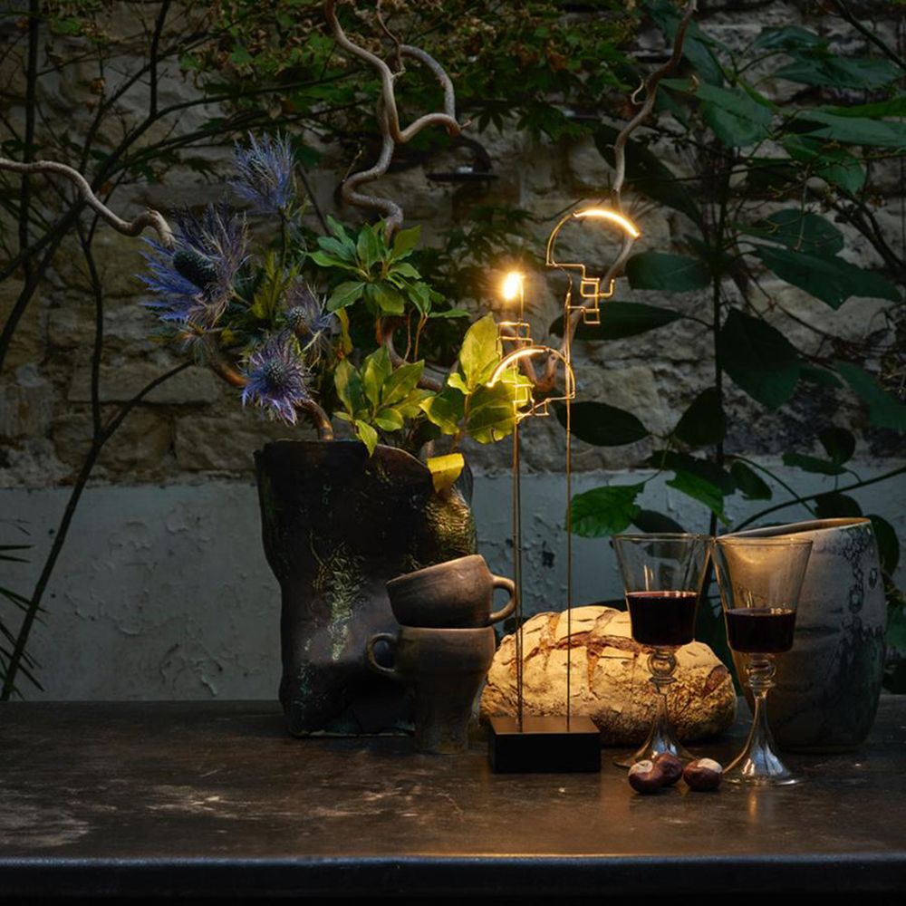 DCWéditions Boucle LED Akku Tischlampe schwarz Gold auf einem Tisch mit Pflanzen und Gläsern.