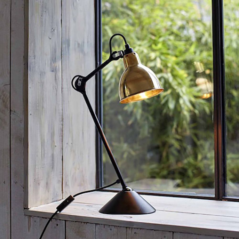 DCWéditions Gras N°205 Tischlampe mit schwenkbarem goldenen Schirm und schwarzem Gestell am Fenster.