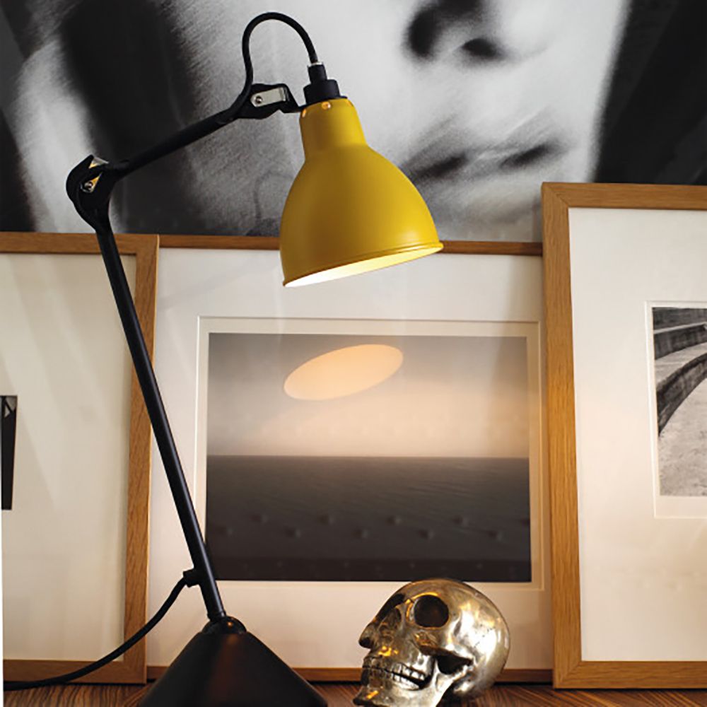 DCWéditions Gras N°205 Tischlampe in gelb, schwenkbar, dekorativ auf einem Tisch, umgeben von Bildern und einer Skulptur.
