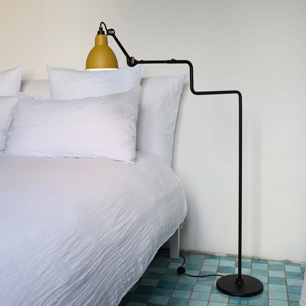 DCWéditions Gras N°411 Stehlampe mit drehbarem Schirm in schwarz und gelb neben einem Bett.