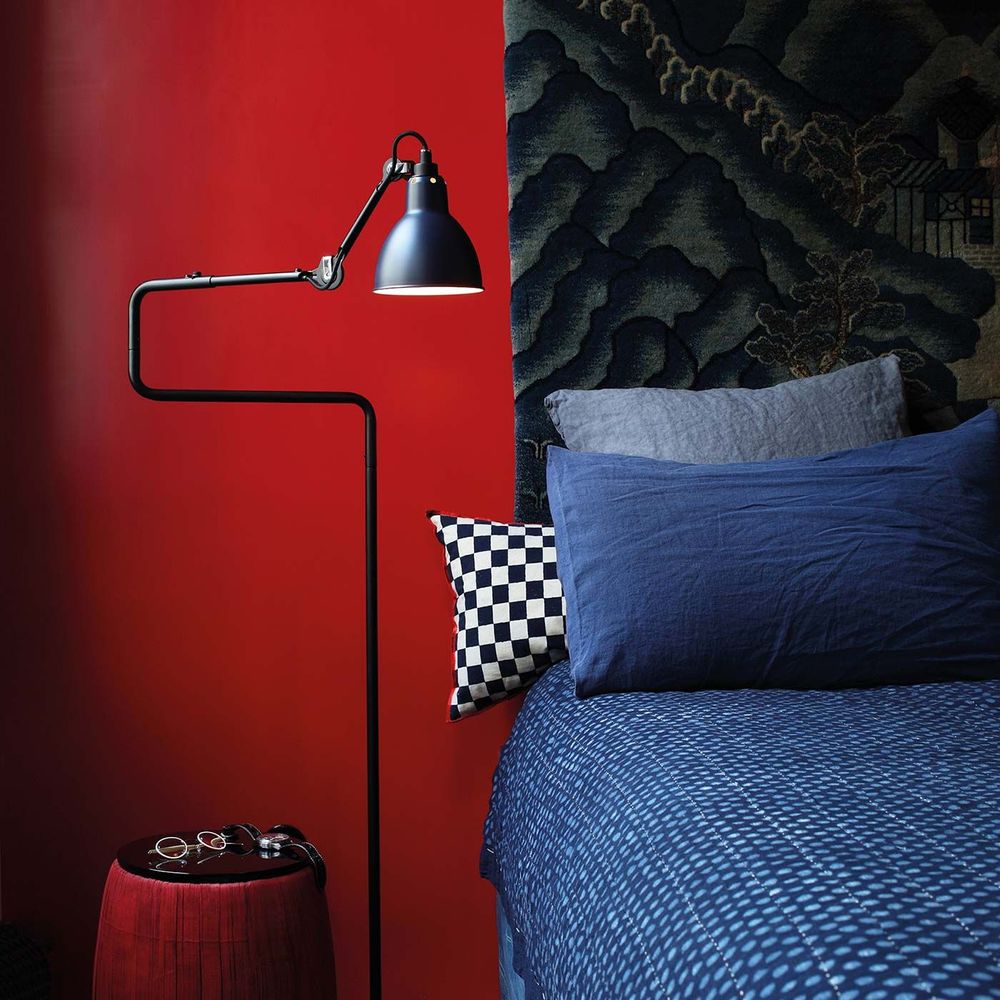 DCWéditions Gras N°411 Stehlampe mit drehbarem Schirm neben einem Bett mit blauen Kissen auf rotem Hintergrund.