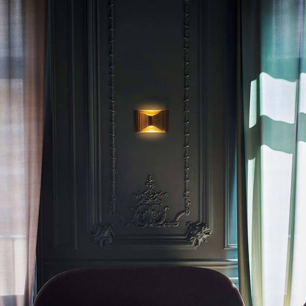 DCWéditions Respiro LED Wandleuchte Gold an einer eleganten Wand mit Vorhängen.