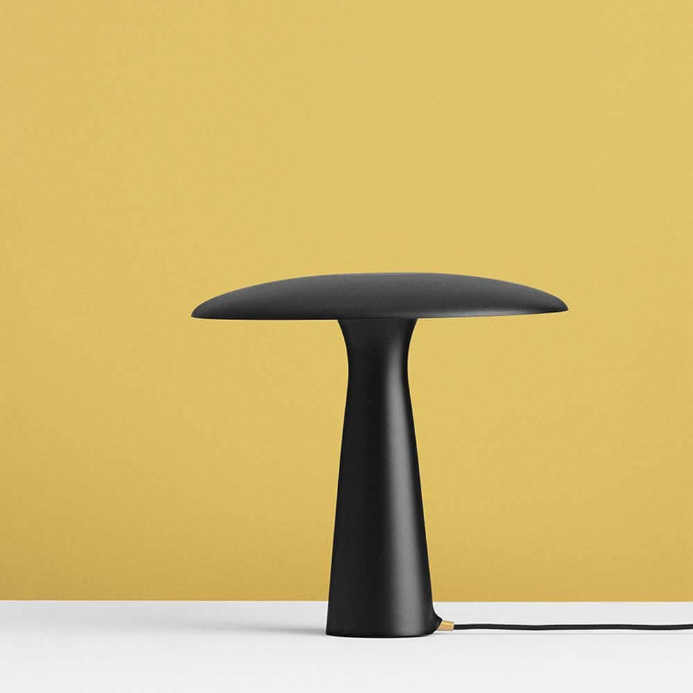 Design Tischlampe Shelter Ø 41cm Schwarz auf weißem Tisch vor gelben Hintergrund.