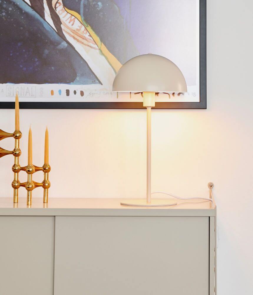 Ellen Tischleuchte Beige auf einem Sideboard mit goldenem Dekor und Kunstwerk im Hintergrund.