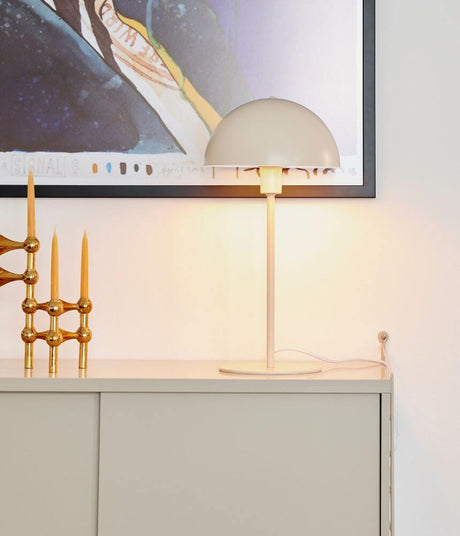 Lampe de table Ellen Beige sur un buffet avec une décoration dorée et une œuvre d'art en arrière-plan.
