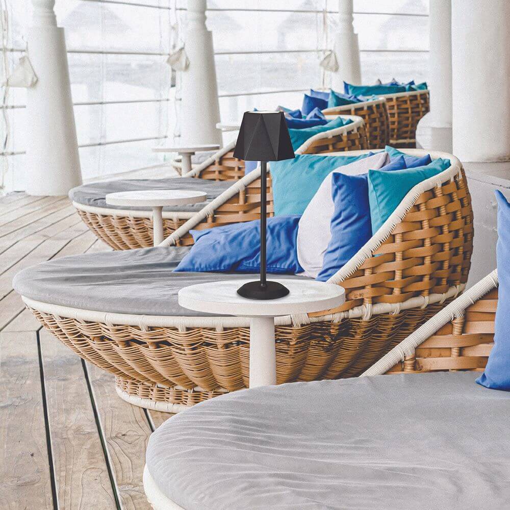 Katy LED Akku-Tischleuchte Metall IP54 auf einem Tisch neben einem Korbsessel mit blauen Kissen.