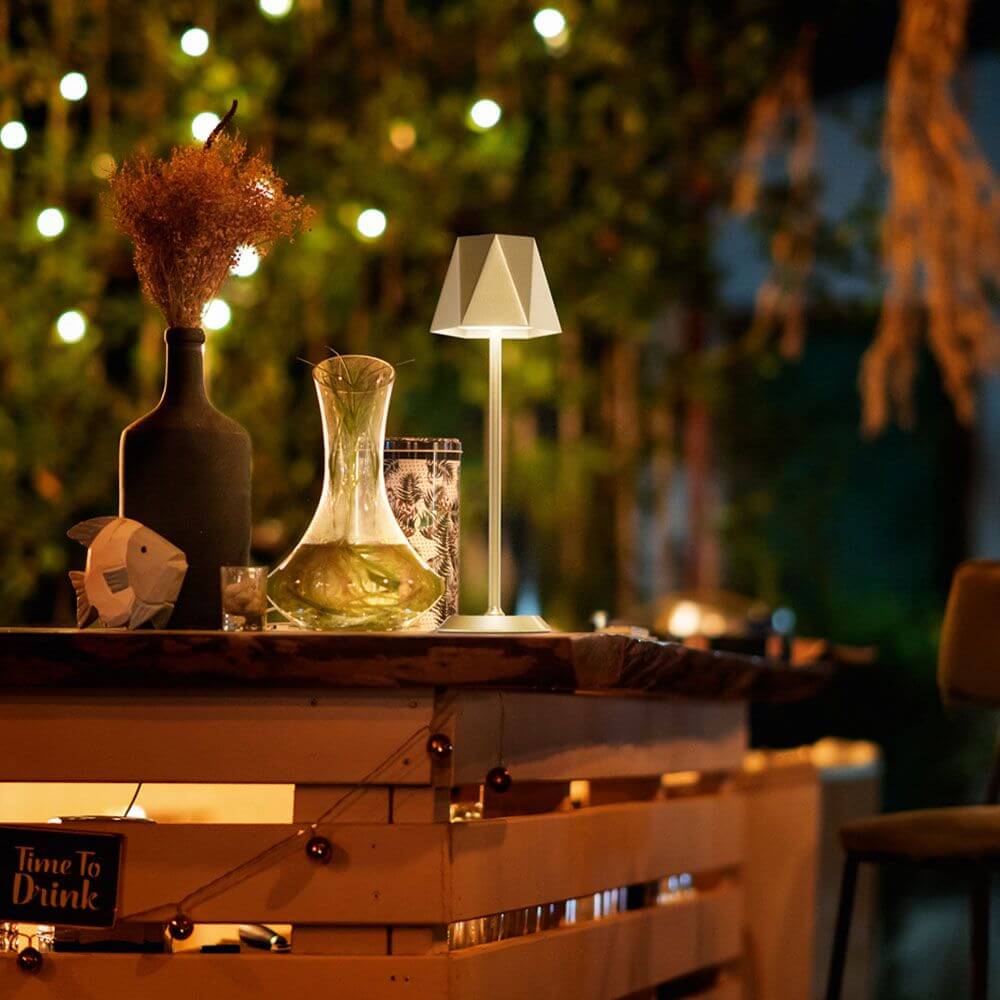 Katy LED Akku-Tischleuchte Metall IP54 elegant beleuchtet auf einem Tisch mit einem Wasserkrug und Pflanzen.