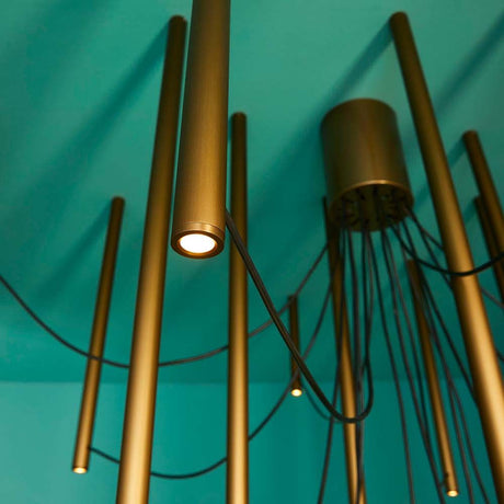 Fabbian Ari LED 16-light pendant light with golden tubes and black cables on a blue wall.