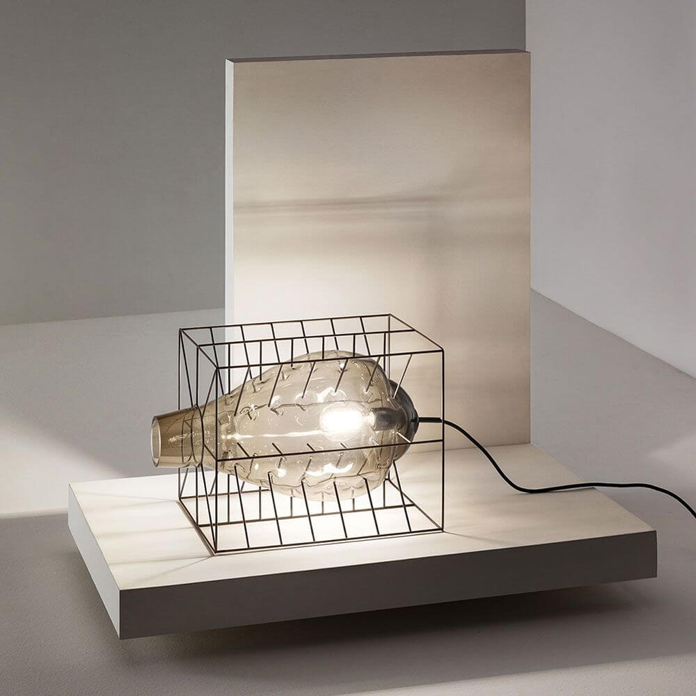 Attimo Tischlampe Muranoglas im Käfig auf einem minimalistischen Tisch mit Lichtscheinen.