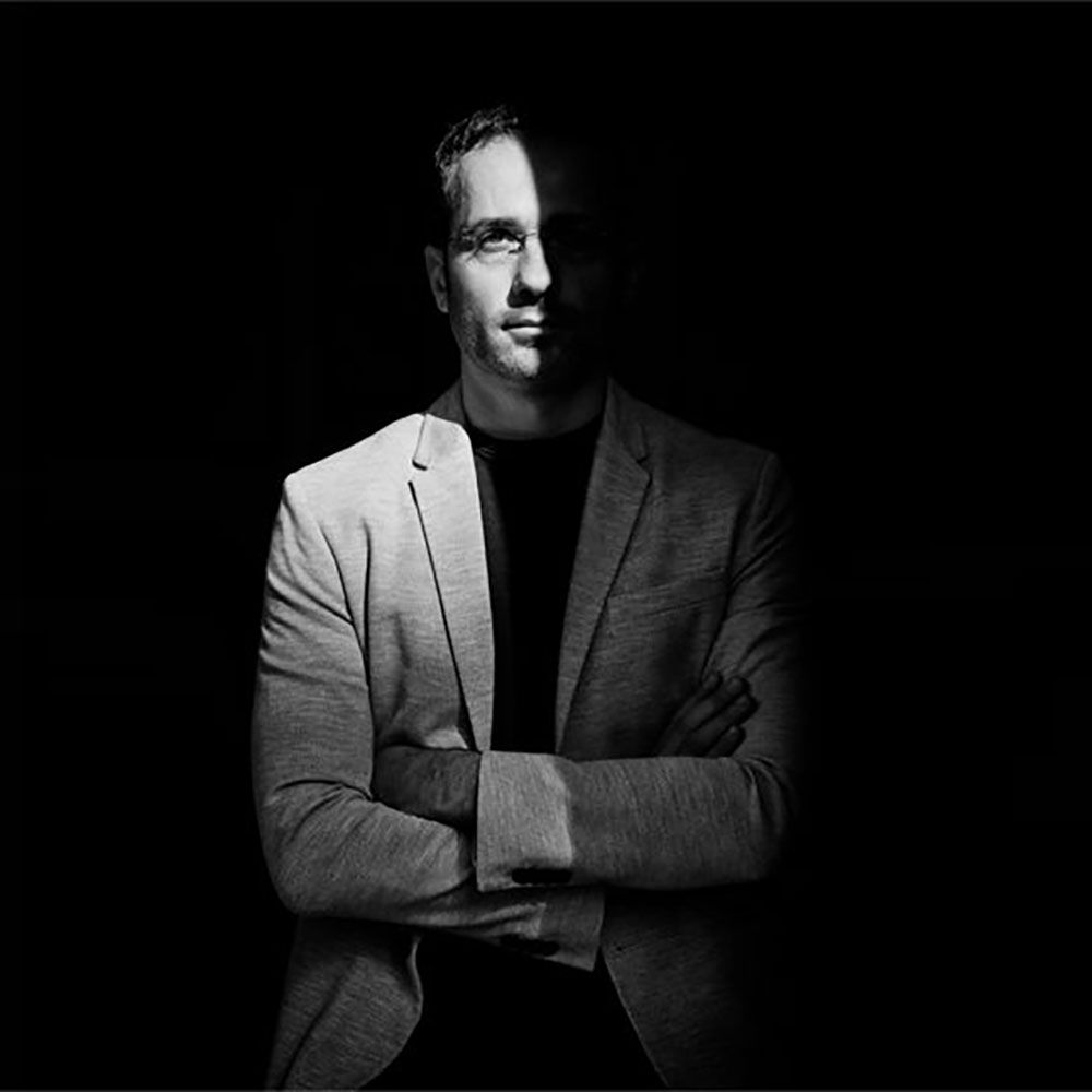 Schwarz-weiße Portraitaufnahme eines Mannes in einem Blazer mit verschränkten Armen im Dunkeln.