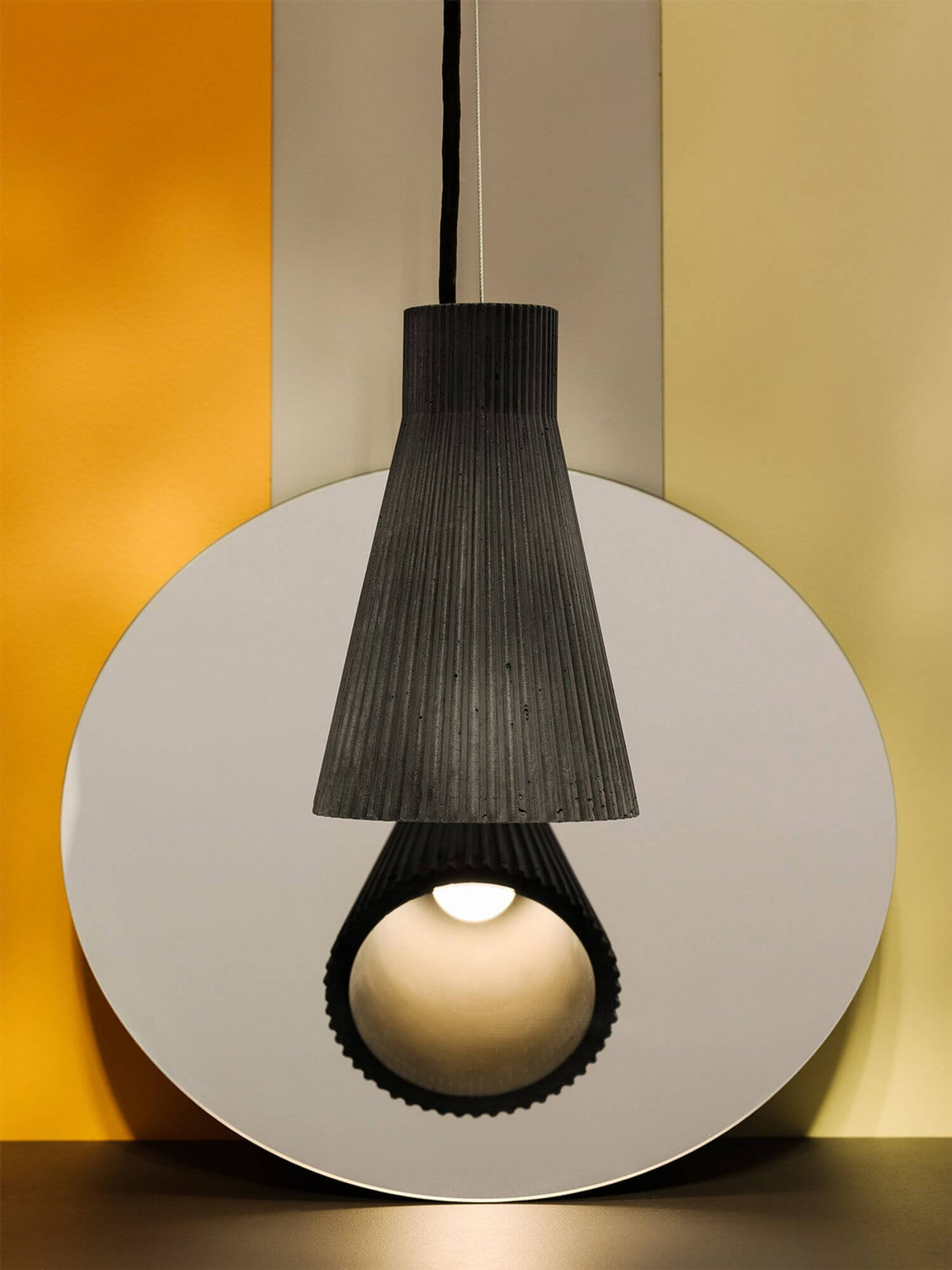 Gantlights S1 Beton Pendelleuchte geriffelt hängt über einem runden Spiegel, betont durch Farben im Hintergrund.