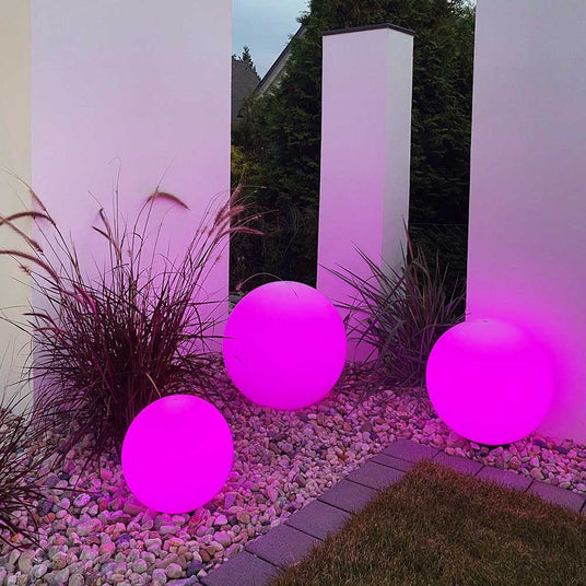 Drei leuchtende rosa Glaskugeln im Garten zwischen dekorativen Pflanzen und weißen Wänden.