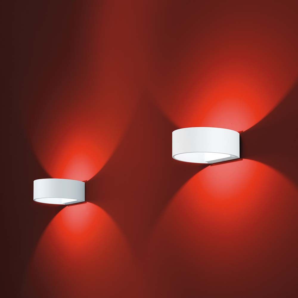 Zwei LED Wandlampen Fosca Weiß an einer roten Wand, die warmes Licht ausstrahlen.