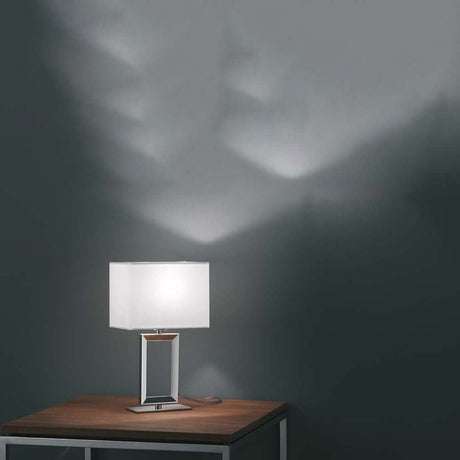 Bordlampe Enna 2 i blank nikkel på et bord med mykt lys og skyggespill på veggen.
