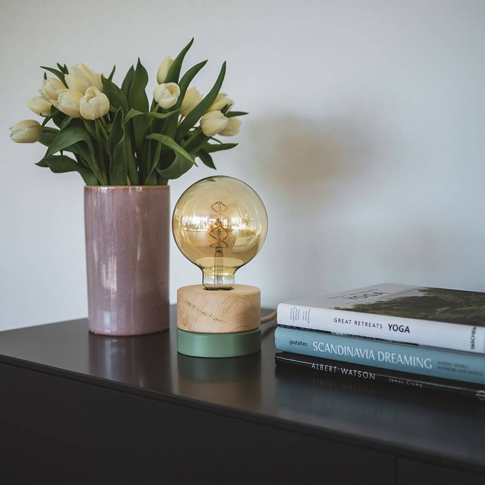 Holz Tischlampe Ø 11cm in 3 Holzarten neben Vase mit Blumen und Bücherstapel auf einem Sideboard.